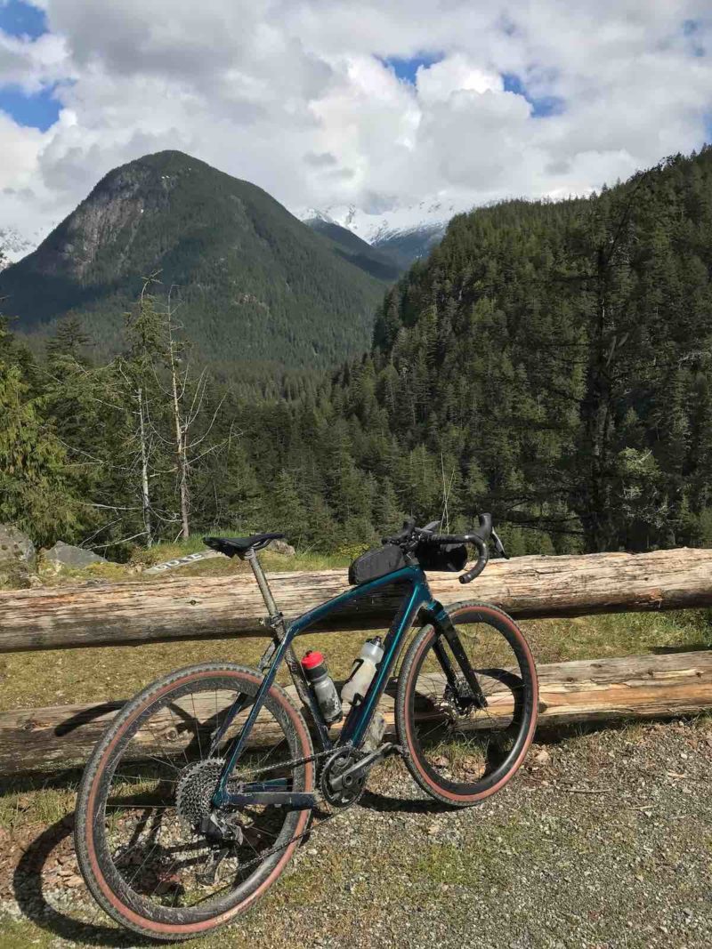 bikerumor pic of the day a mountain bike is next to a log fence overlooking mountain peaks covered in pine trees, the sun is bright and there are fluffy white clouds in the distance.
