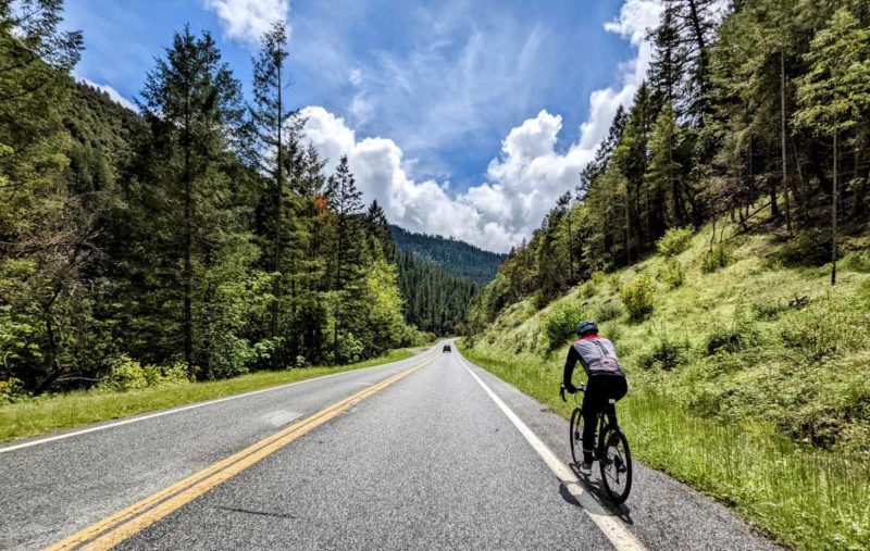 bikerumor pic of the day a cyclist ride alongside a road with high mountains on either side covered in lush green grasses and pine trees, the sky has high fluffy white clouds and the sun is bright and high over the cyclist.