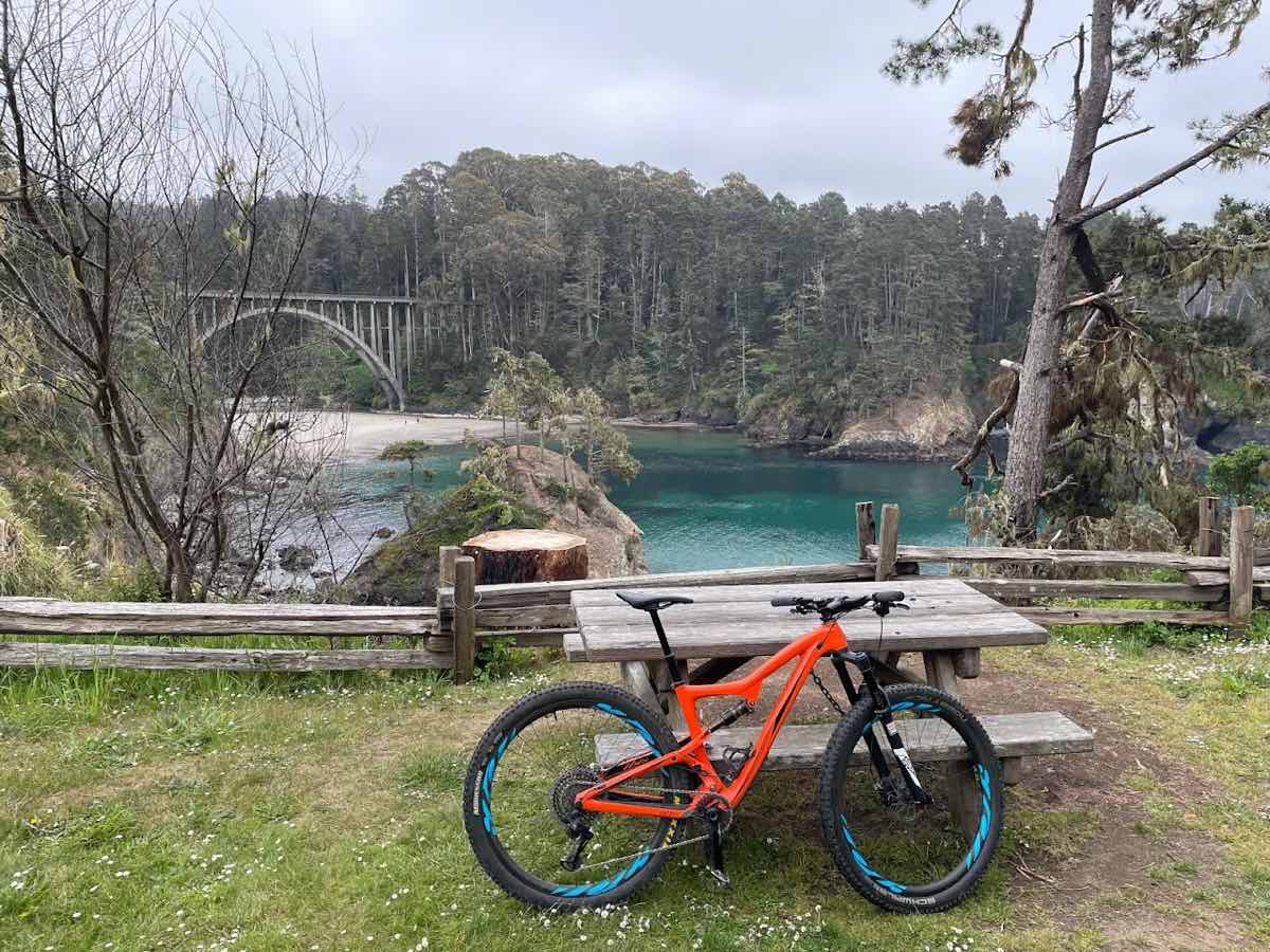 bikerumor pic of the day a red mountain bike leans against a wood picnic table on a grassy area overlooking a blue lake or river with a train bridge spanning over it