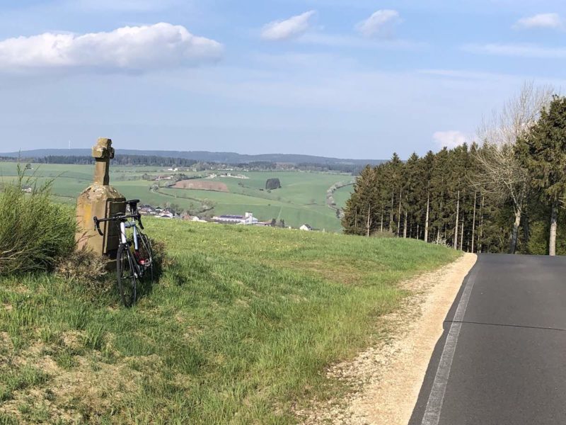 bikerumor pic of the day a bicycle leans against a stone marker with a cross on the top, there is a paved road to the right that has a grove of trees along one side and fields for as far as the eye can see on the other side, the day is sunny and there are some fluffy clouds in the sky.