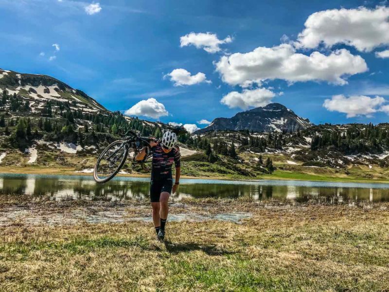 bikerumor pic of the day a cyclist carries their bike walking in a grassy field away from a lake that is at the base of some large rocky mountains, there is snow and some pine groves on the mountains, the sky is bright and there are some sparce fluffy clouds.