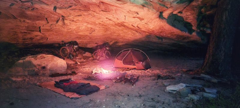 bikerumor pic of the day a campsite inside a cave with a tent, bicycles for bikepacking and a small fire the glow of the fire reflects off the inside of the cave creating a warm glow.