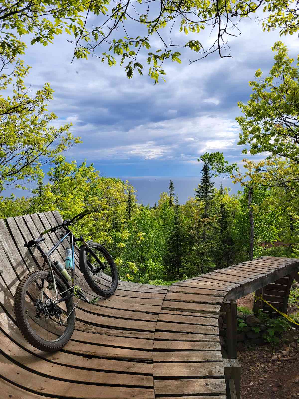 bikerumor pic of the day a wooden mountain bike trail bridge ramp has a mountain bike leaning against it, in the distance are pine trees and a harbor with a freighter that is very far away, there are clouds in the sky that are dark but it is sunny over the trees and wood trail.