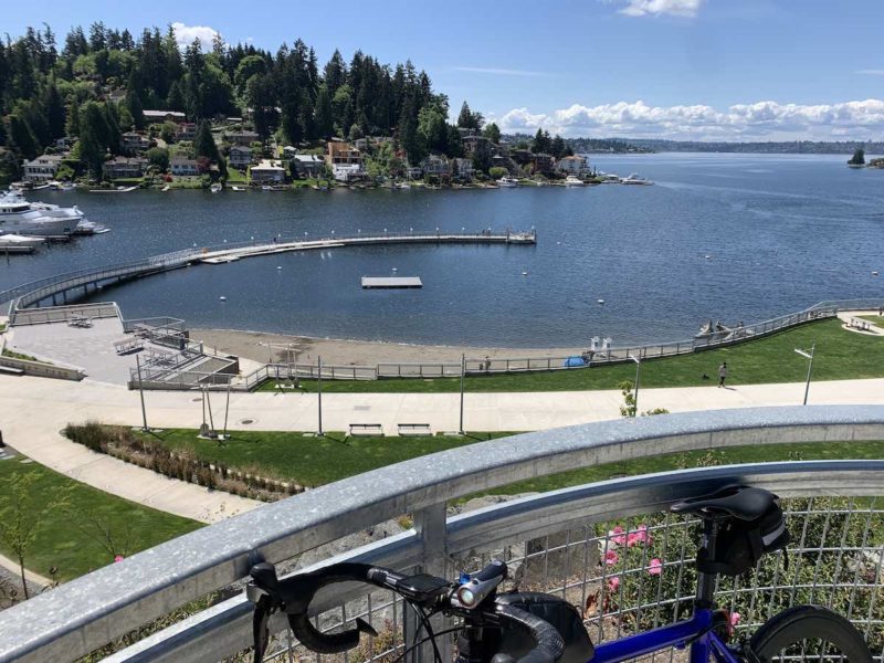bikerumor pic o the day a road bicycle leans against a metal railing in the foreground, in the distance is a small bay with a beach on one side and a small peninsula covered in trees and houses along the waterfront, the sky is clear and there are fluffy clouds on the horizon.