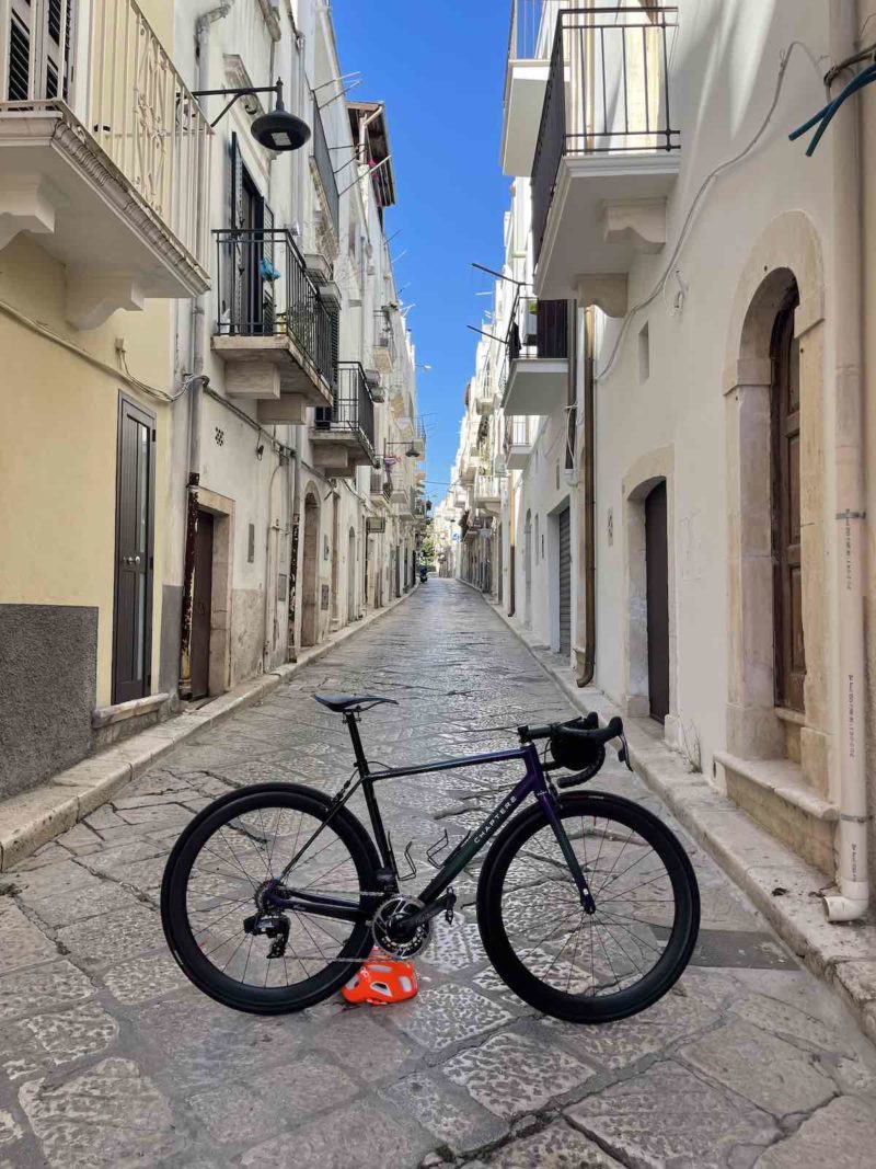 bikerumor pic of the day road bike leaning against a red helmet that's been placed on the ground in the middle of a small thoroughfare between cream colored buildings with clear bright blue sky above.