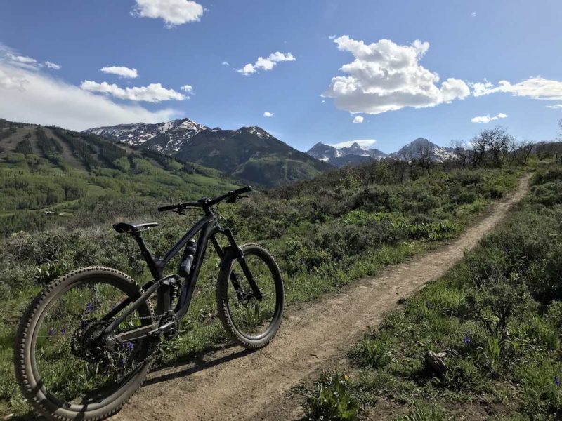 bikerumor pic of the day a mountain bike is aimed away from the camera along a narrow dirt trail heading into the brush surrounded by low mountain range and blue skies with fluffy white clouds here and there.
