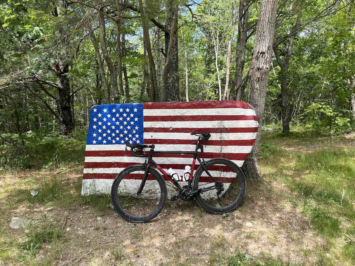 bikerumor pic of the day a bike leans against a large flat rock that has the american flag painted on it, the rock is in a grassy clearing near a section of leafy green trees.