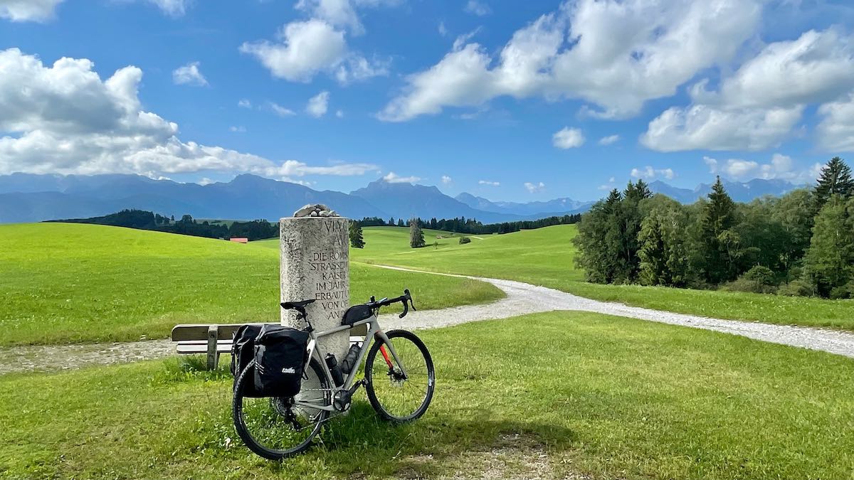 bikerumor pic of the day a bicycle leans against a stone marker on the via claudia augusta, there is a path along short green grass and trees off to one side and the distance the sky is bright and there are a few fluffy white clouds.