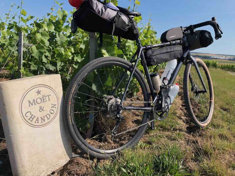 bikerumor pic of the day a bikepacking trip along the Via Francigena into the champagne region the bike with its packs is leaning against a marker that reads moet & chandon and it is next to a grape field the sun is high and it is very bright the sky is clear
