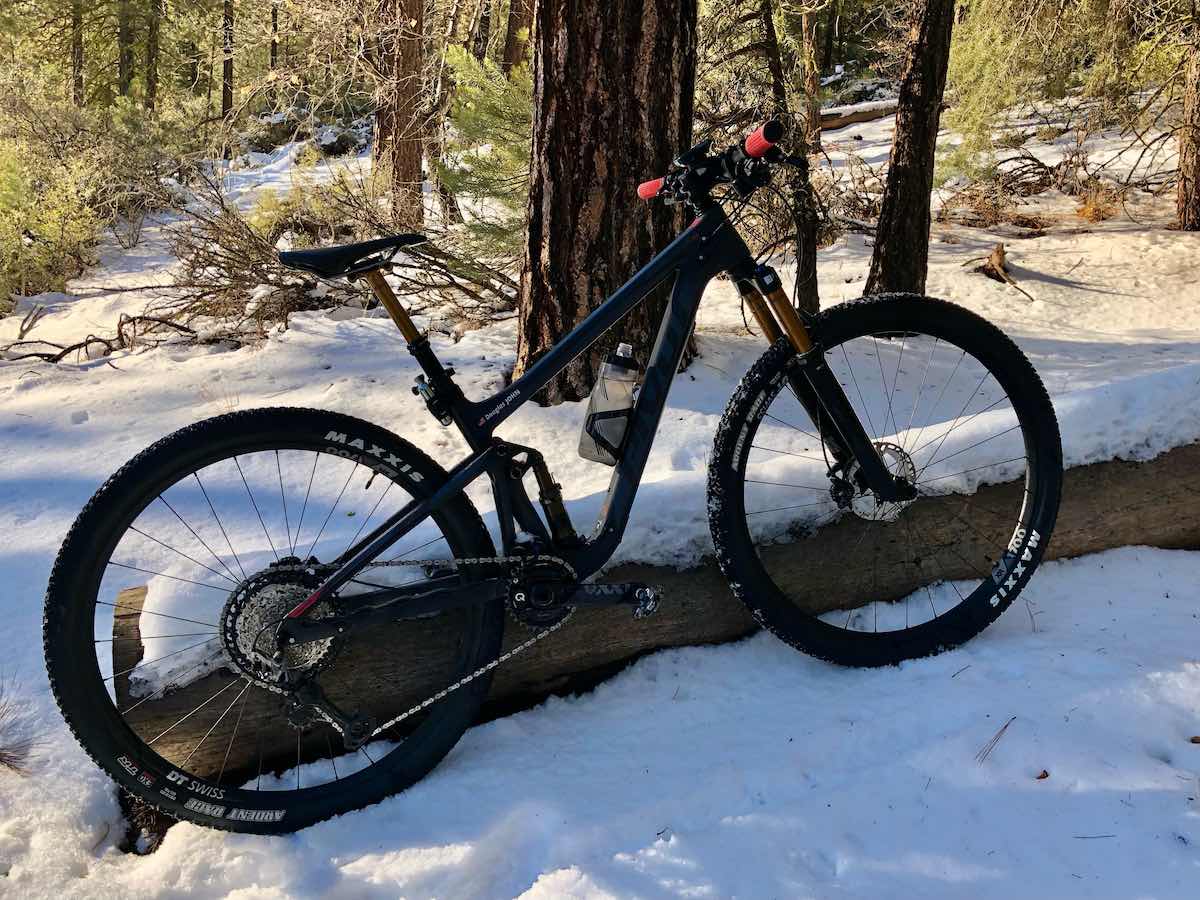 bikerumor pic of the day a mountain bike is leaning against a log in about 4 inches of snow, it is sunny out and there are patches of green trees in the background