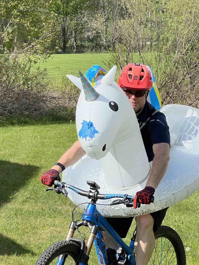 bikerumor pic of the day a mountain biker is riding in a grassy field towards the camera, besides a helmet and gloves they are wearing an inflatable inner tube around their waist that is in the shape of a white unicorn.