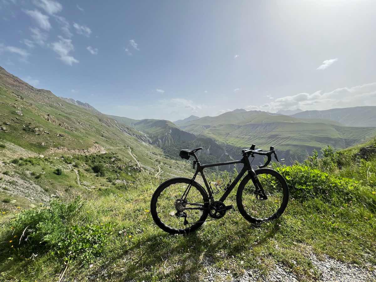 bikerumor pic of the day a black road bicycle sits at the top of the Alpe d’Huez on a green grassy ridge overlooking an expanse of green mountains the sun is bright and off to the side