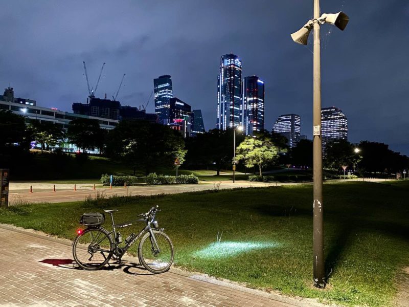 bikerumor pic of the day a vado bicycle is parked on a brick trail with the skyline of Seoul in the distance it is night and the trail is lit up and the city has many bright lights.