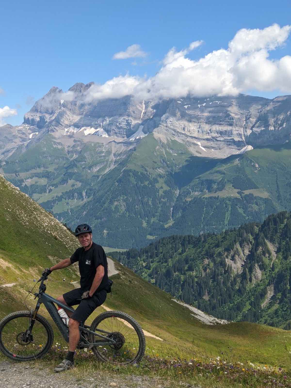 bikerumor pic of the day a cyclist sits on their bike on the edge of a mountain pass overlooking the alps, it is a sunny day.