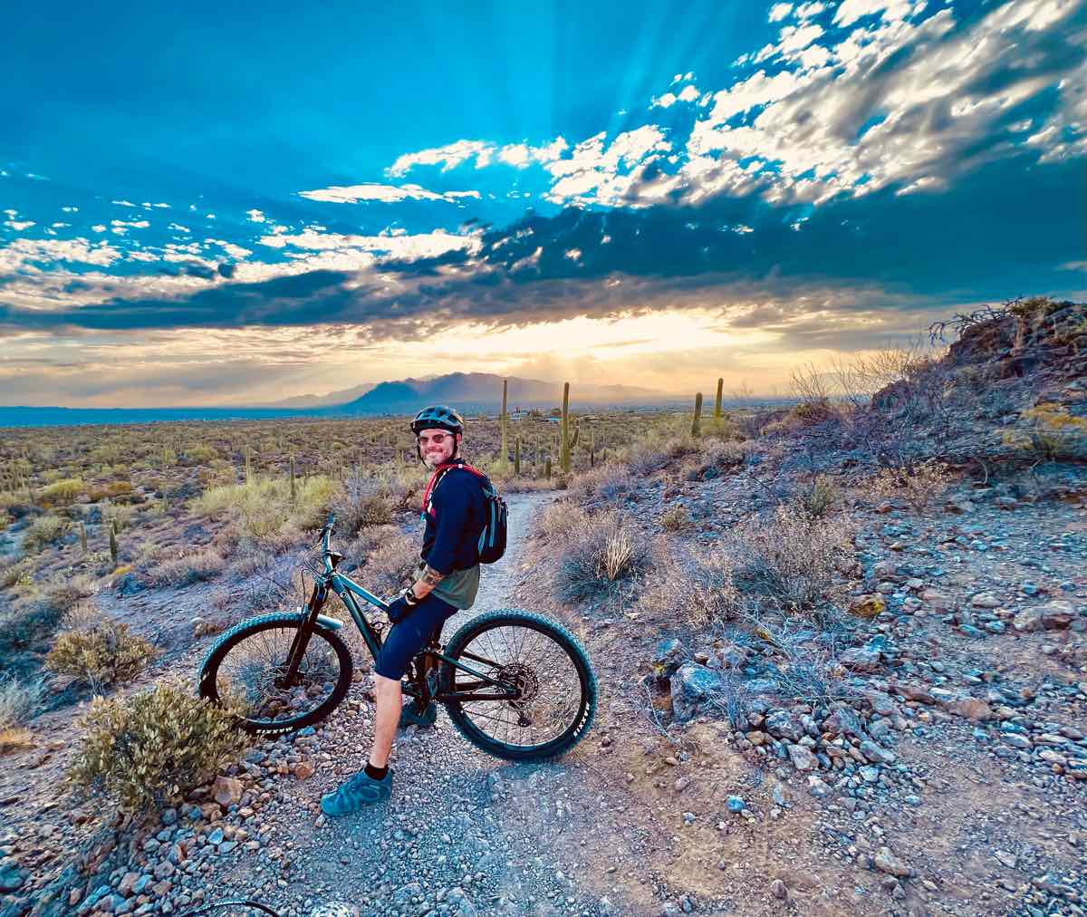 bikerumor pic of the day a mountain biker sits on their bicycle on a rocky dirt trail in arizona there is scrub brush on either side of the trail and a low mountain in the distance the sun is very low and is blocked by a series of clouds along the horizon creating rays that shoot up into the sky.