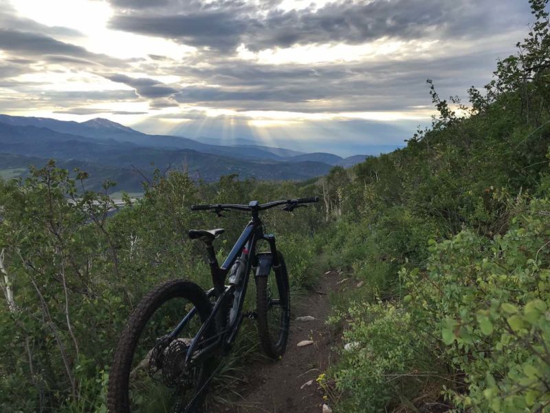 bikerumor pic of the day a mountain bike is on a narrow dirt trail on the side of a mountain surrounded by short green brush the clouds in the sky are parting to let rays of sunshine out in the distance and it looks like has just rained.