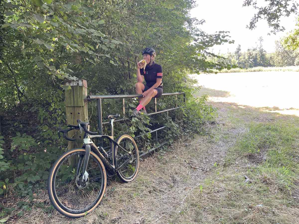 bikerumor pic of the day a gravel cyclist perches on a gate eating a snack as their bike rests against the gate next to a path by a field.