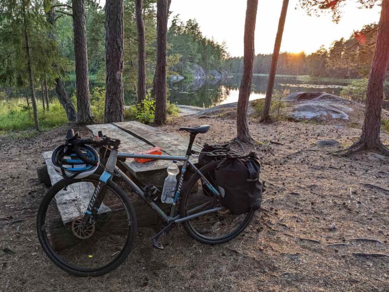 bikerumor pic of the day a bicycle with packs leans against a wood picnic table in a clearing by a lake, the sun is low and bright over the horizon.
