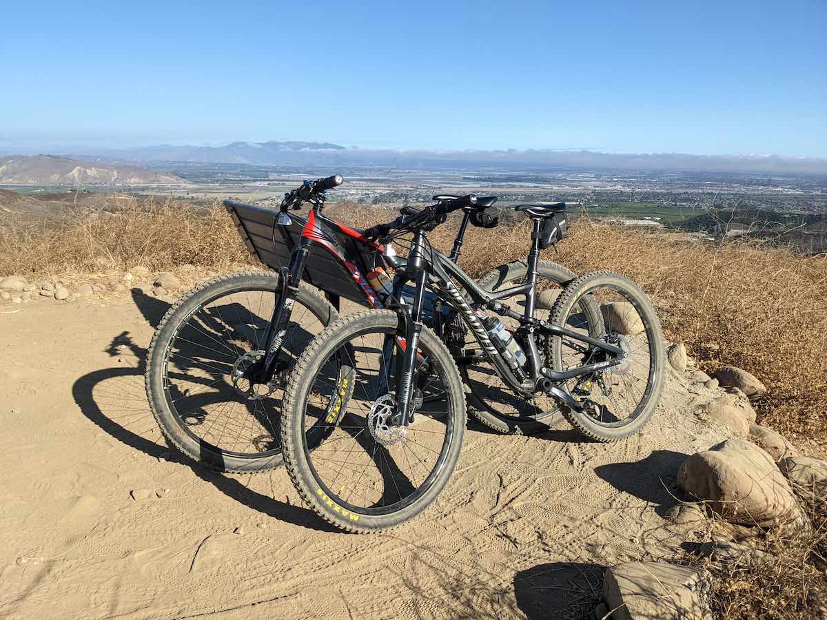 bikerumor pic of the day two mountain bikes lean against a bench on a dirt overlook with a view of a large city in a valley below there is a low cloud formation at the horizon and clear skies above