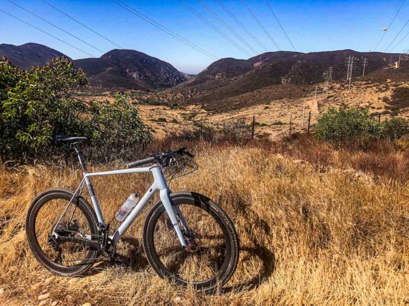 bikerumor pic of the day a mountain bike is in golden field of grass with small cruz trees and low hills in the distance, the sky is clear and blue and contrasts well with the brown green hues of the land.