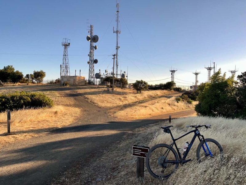 bikerumor pic of the day a mountain bike is posed near a dirt trail that leads up to a peak that is covered in telecommunication towers and dishes, the sun is low and the sky is clear.