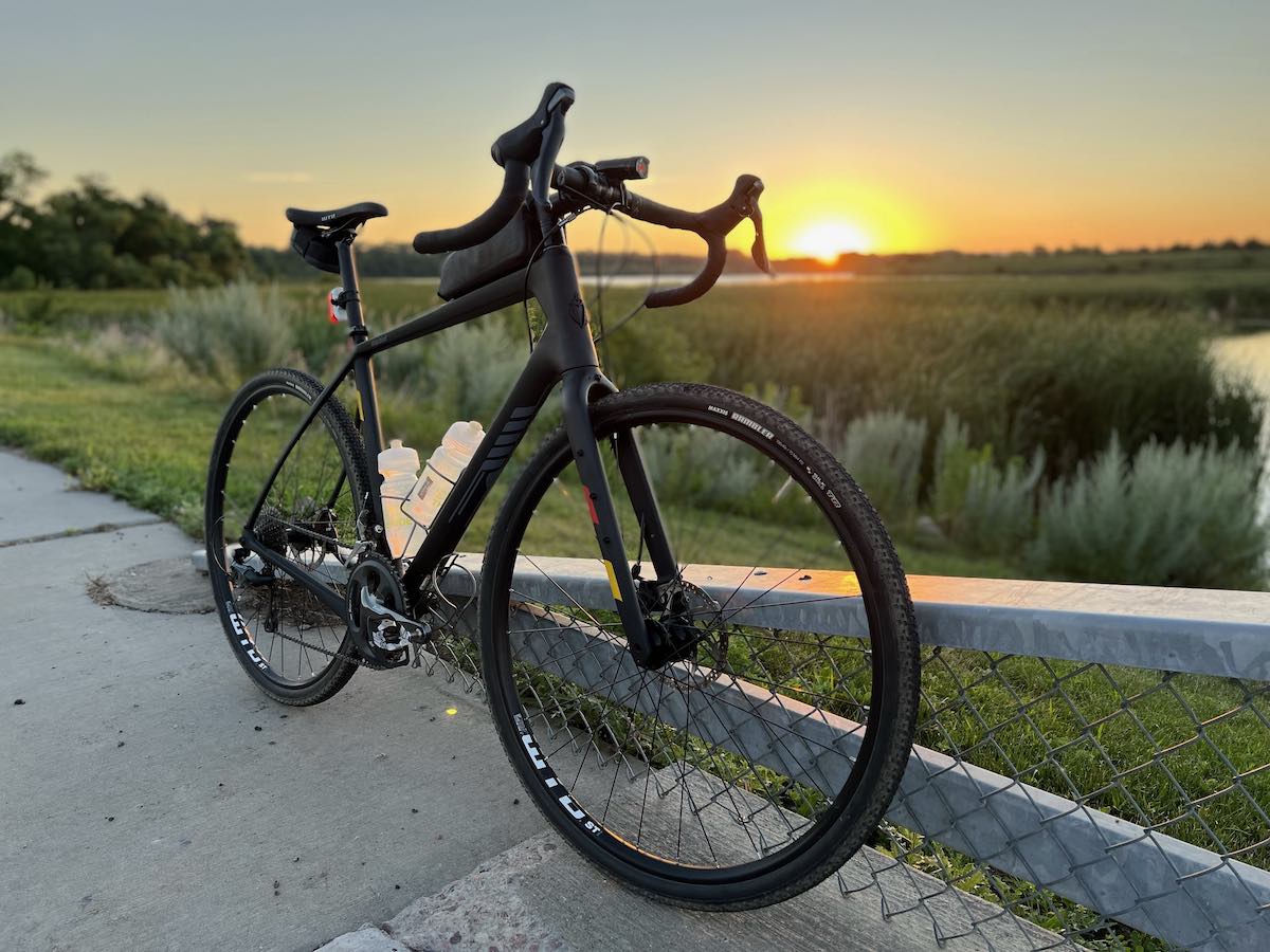 bikerumor pic of the day a bicycle is on the side of a road at sunrise, there is a lake with tall grass surrounding it and the sunrise is on the horizon behind the bike, it is a clear morning and the sun is casting a golden glow over the sky.
