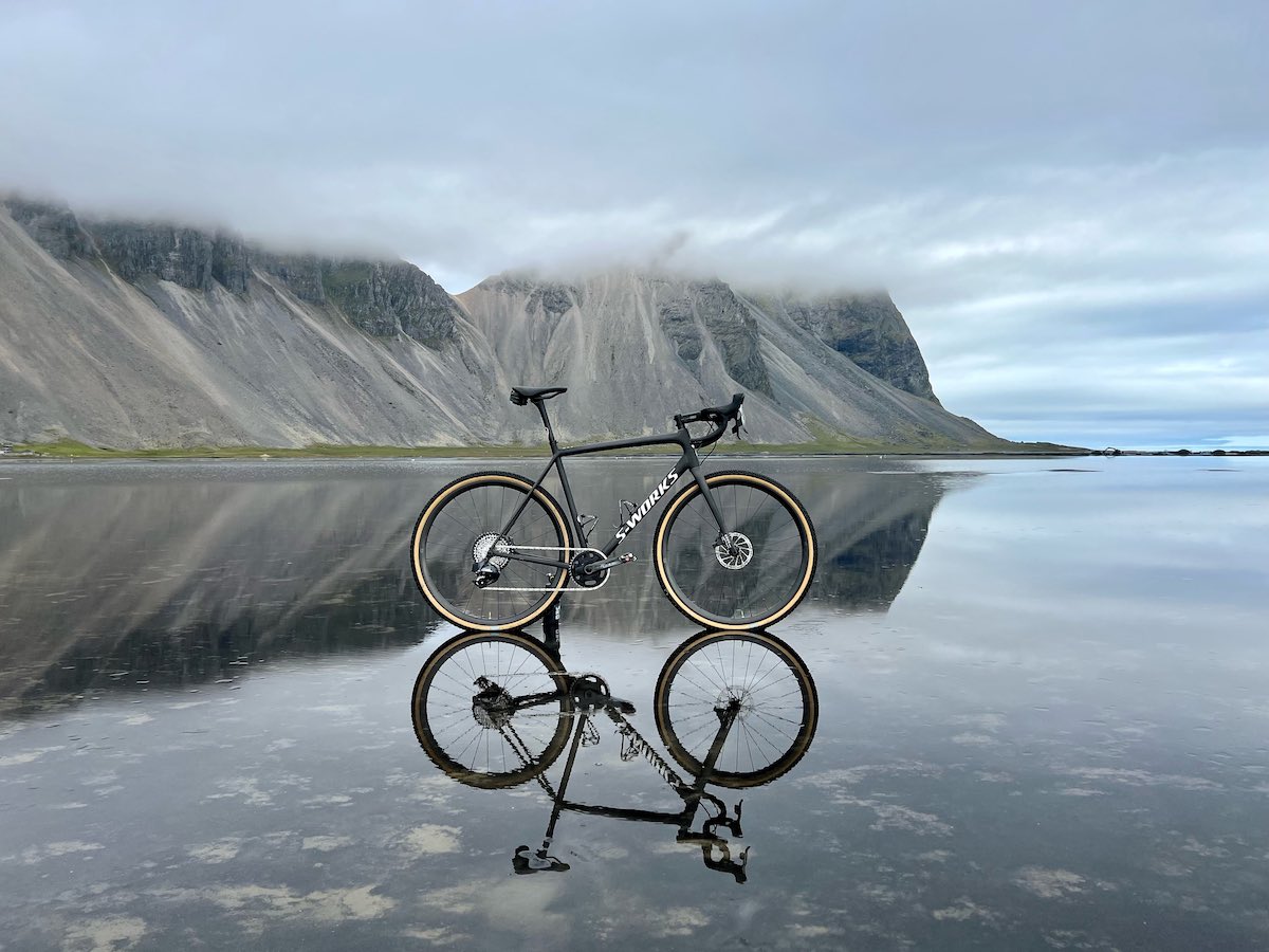 bikerumor pic of the day a specialized gravel bike looks like it is levitating on top of smooth water surrounded by steep cliffs of the iceland shoreline, the sky is cloudy and the water looks like glass.