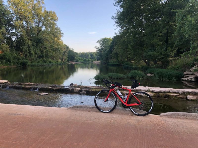 bikerumor pic of the day a red bicycle is on a path overlooking a water bridge surrounded by leafy green trees.