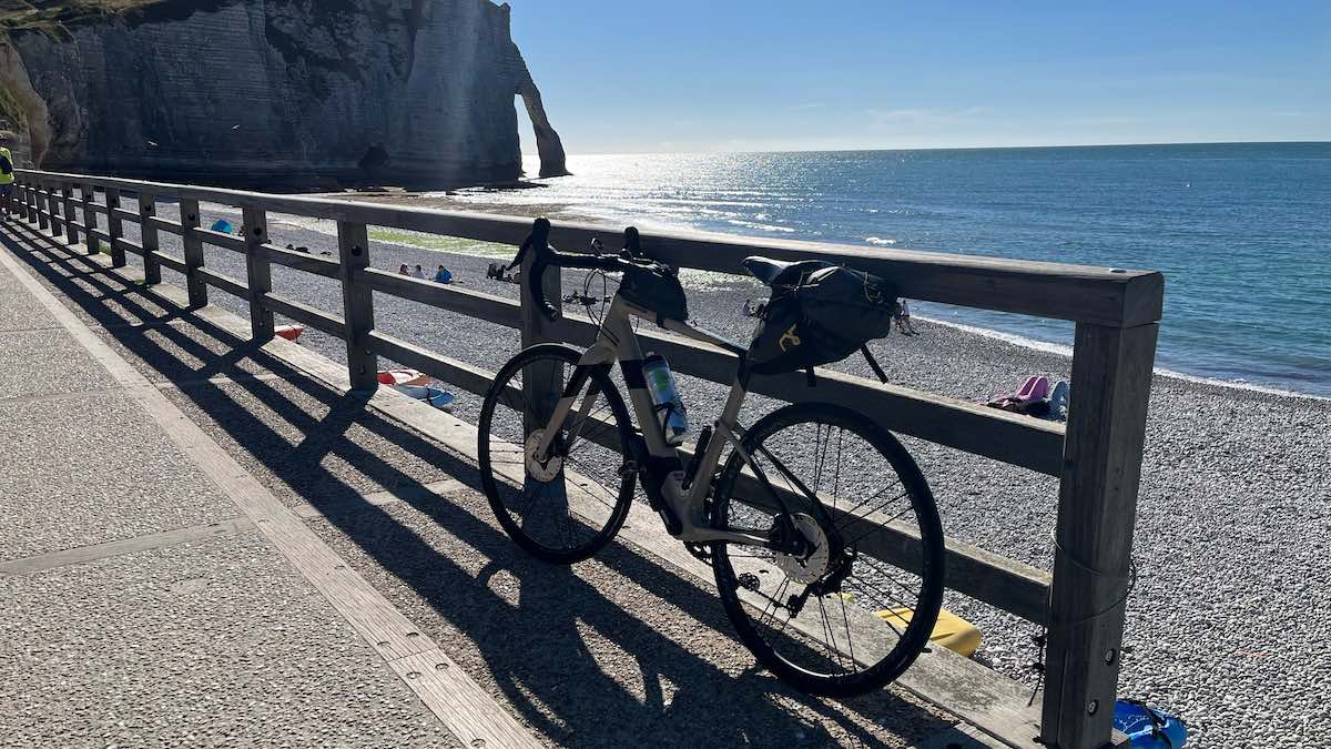 bikerumor pic of the day a road bicycle leans against a wood railing alongside a road that has a small beach on one side, there are small groups of people on the beach and the sun is shining very bright on the still water.