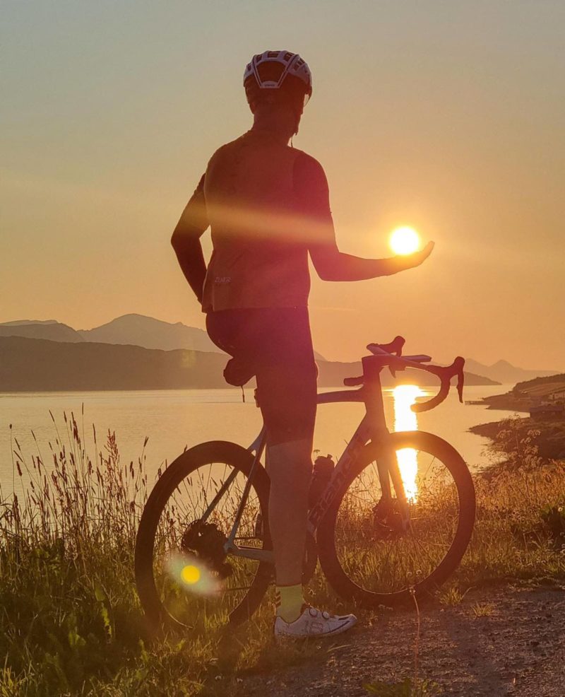 bikerumor pic of the day a person on a bike is on the edge of a body of water looking out over the water towards the setting sun, they stand with their arm outstretched and the perspective makes it look as though the sun is sitting on the palm of their hand.