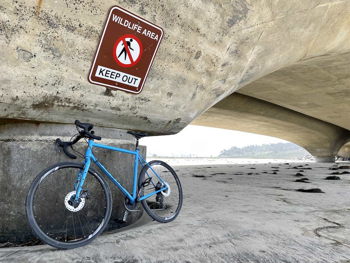 bikerumor pic of the day a bicycle leans against a concrete base of a bridge on a sand packed beach.