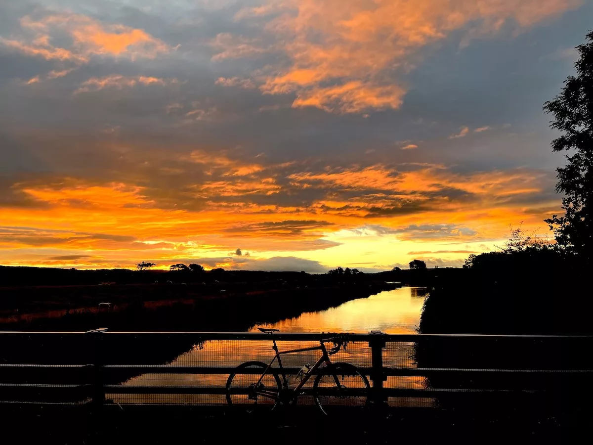 bikerumor pic of the day a bicycle leans against a metal railing over a small river, the sun is rising in the distance and creating a very bright orange glow over the dark land and clouds.