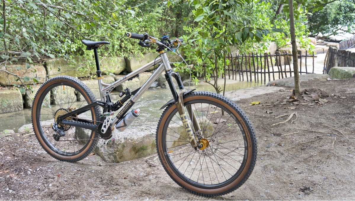 bikerumor pic of the day a mountain bike is posed in the dirt near a stream bordered with large bricks and tropical trees on the other side.