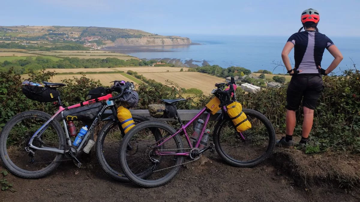 bikerumor pic of the day a cyclist stands next to to bicycles loaded with packs overlooking a low land and bay with fields and a small city.