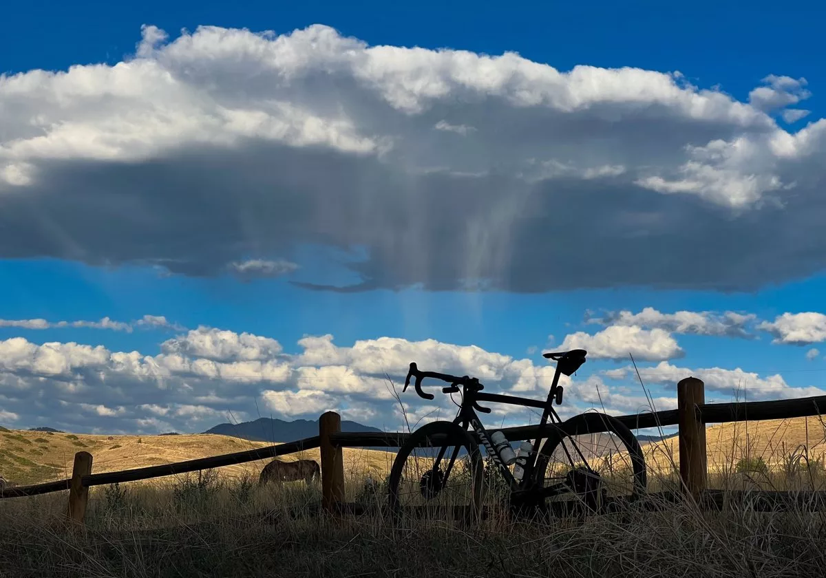 bikerumor pic of the day a bicycle leans against a wooden split-rail fence next to a golden field of grass with a low mountain in the distance and large white fluffy clouds in a bright blue sky