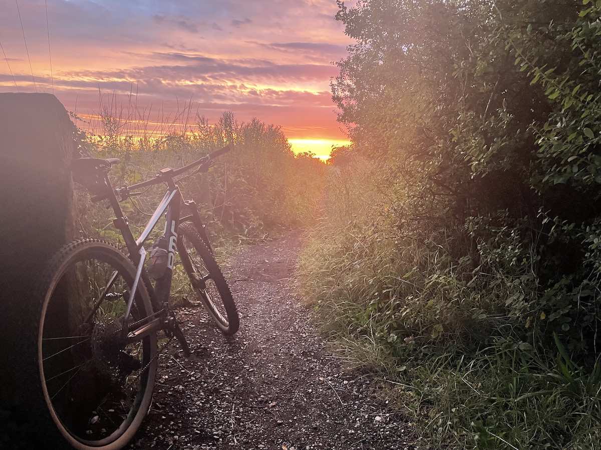 bikerumor pic of the day a mountain bike is to the side of a trail with scrub bushes on either side the sun is setting in the distance and the sky is bright orange and grey and hazy with clouds