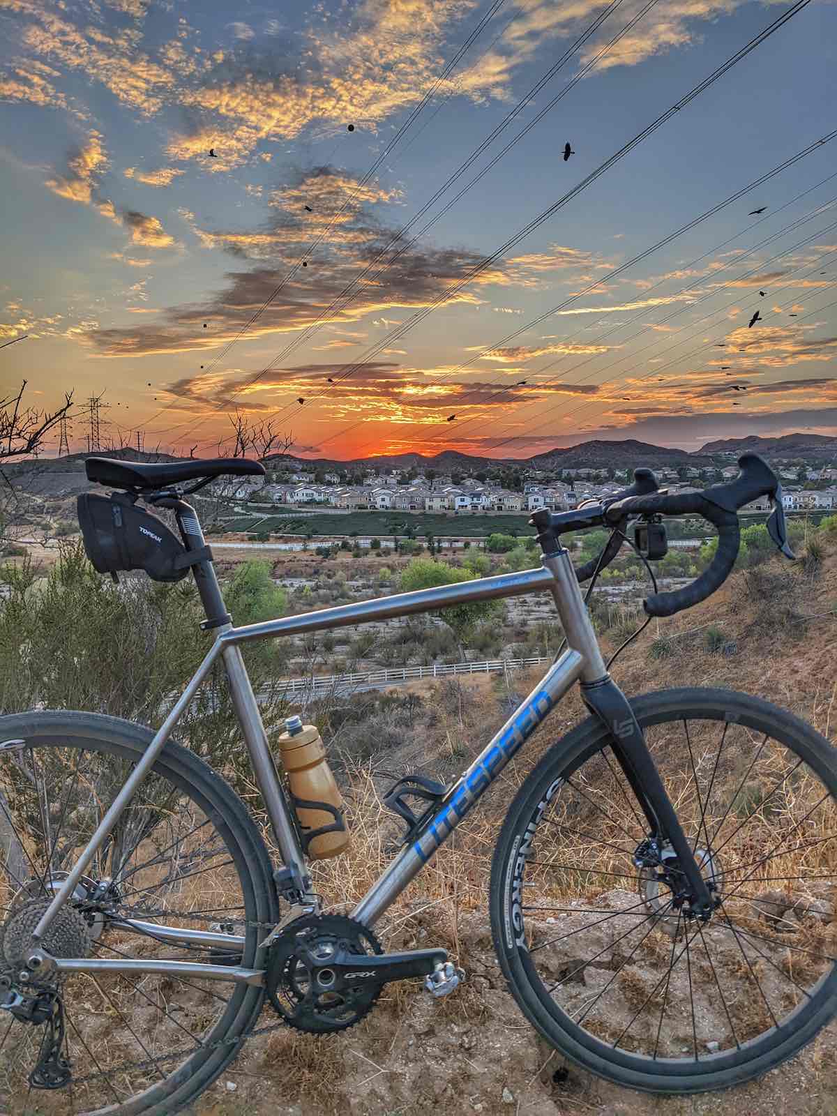 bikerumor pic of the day a titanium bicycle is in the foreground before a desert like field with a city filled with homes in the distance at dusk with the sun just below the horizon.