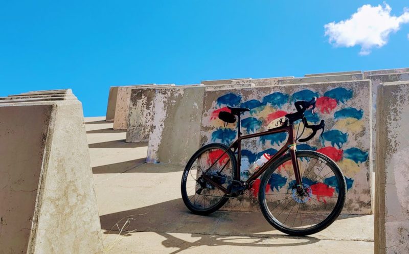 bikerumor pic of the day a gravel bike leans against a spillway structure on the john b waters dam in new mexico, there is graffiti of buffaloes spray painted in red and blue on one of the concrete flanges, and there is one perfect white cloud in the clear blue sky.