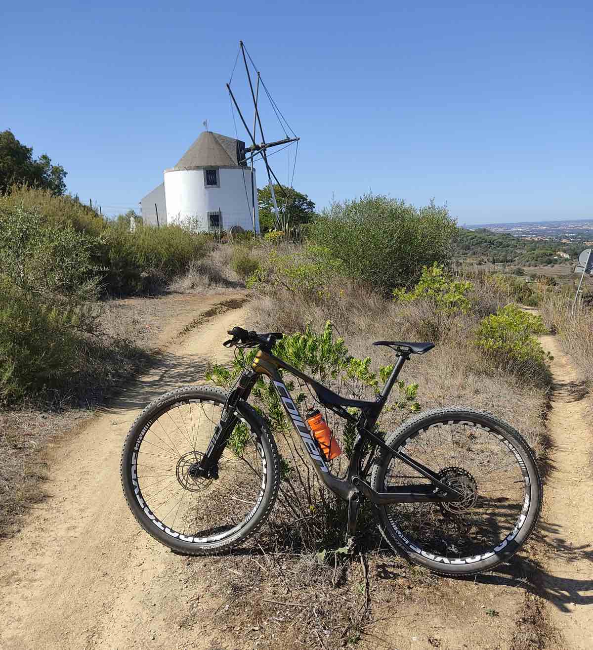 bikerumor pic of the day a mountain bike is on the side of a dirt trail leading up towards a squat windmill there is brush along the trail and the sky is clear and blue