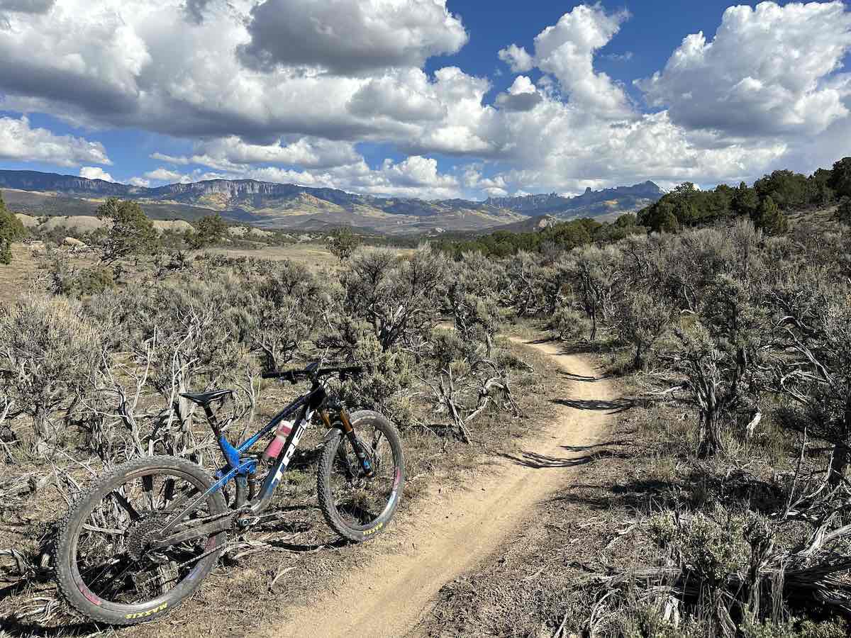 bikerumor pic of the day a mountain bike is beside a dirt single path surrounded by scrub brush in a low valley with mountains in the distance, the sky is dotted with light fluffy clouds and the sun is high and bright.