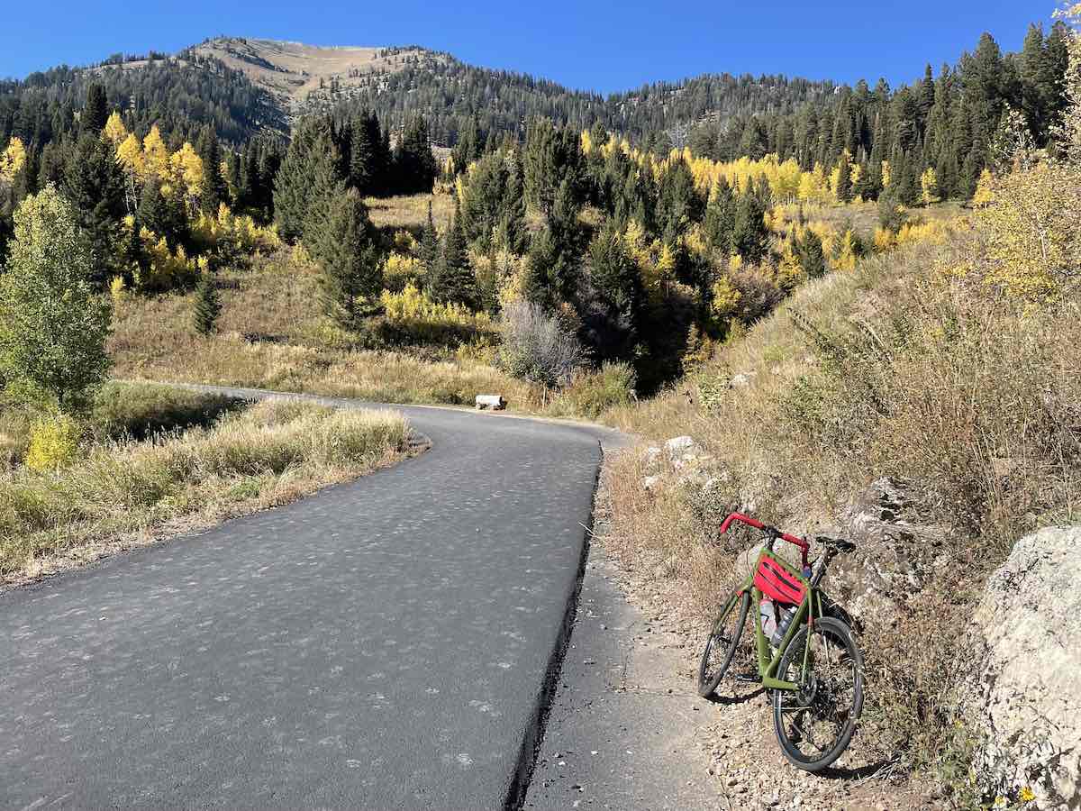 bikerumor pic of the day a road bike leans on a rocky outcrop along a single paved lane going up a mountain covered in pine tree and trees with yellow leaves, there is a red pack on the bicycle that is very bright and stands out among the fall colors.