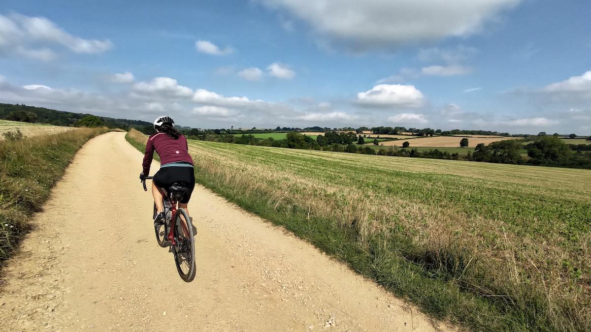 bikerumor pic of the day a cyclist on a dirt road on their lost lane tour surrounded by grass fields on undulating land with sunny sky and dotted with fluffy clouds