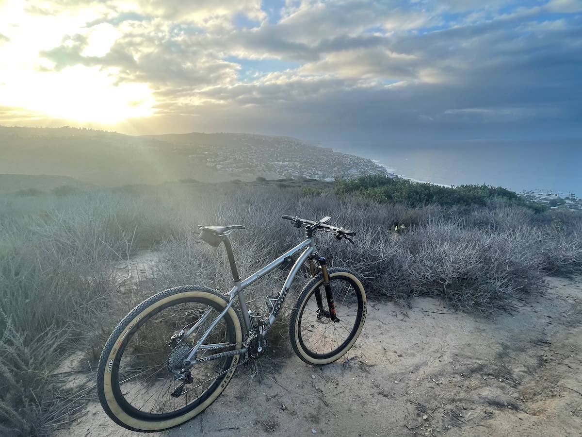Bikerumor pic of the day a bicycle is on a sandy trail amidst scrub brush overlooking a small city near the ocean, the sky i s cloudy and the sun is peeking out towards the horizon