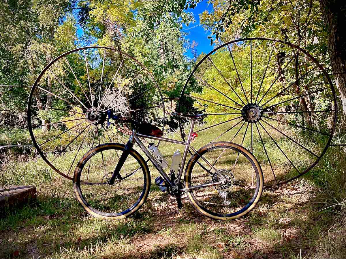 bikerumor pic of the day a gravel bike leans against two large metal wagon wheels that block a road along a dirt trail among trees.