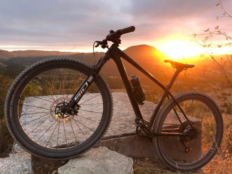bikerumor pic of the day a mountain bike is propped up on a slab of stone with a low landscape behind with the sun rising and a semi cloudy sky.