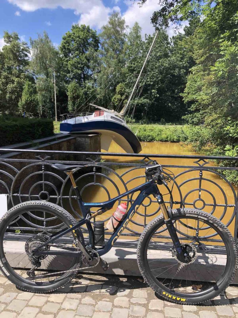 bikerumor pic of the day a bicycle leans against a decorative metal railing alongside a canal, surrounded by trees, a sculpture of a sailboat rests on the side of the canal and looks like it curves almost cartoonish in such a way that it is hugging the wall it is placed on.