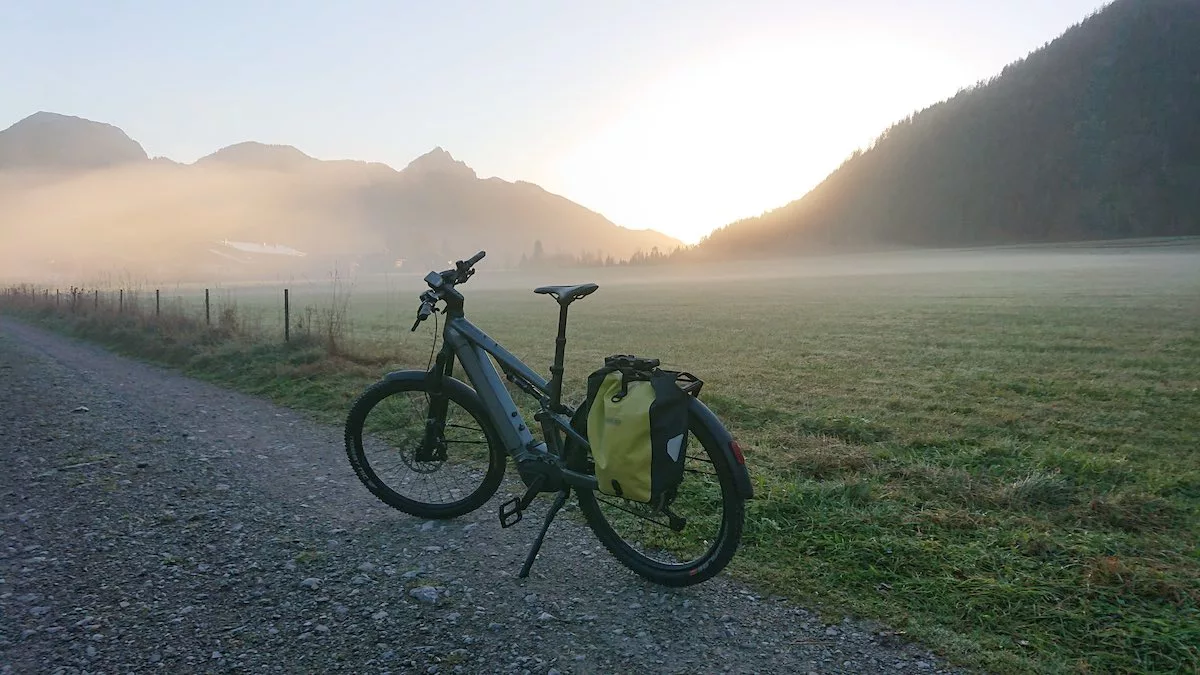 bikerumor pic of the day an e-bike is parked on a gravel path next to a field at the bottom of a mountain as the sun peeks over the horizon and fog dissipates off the field.