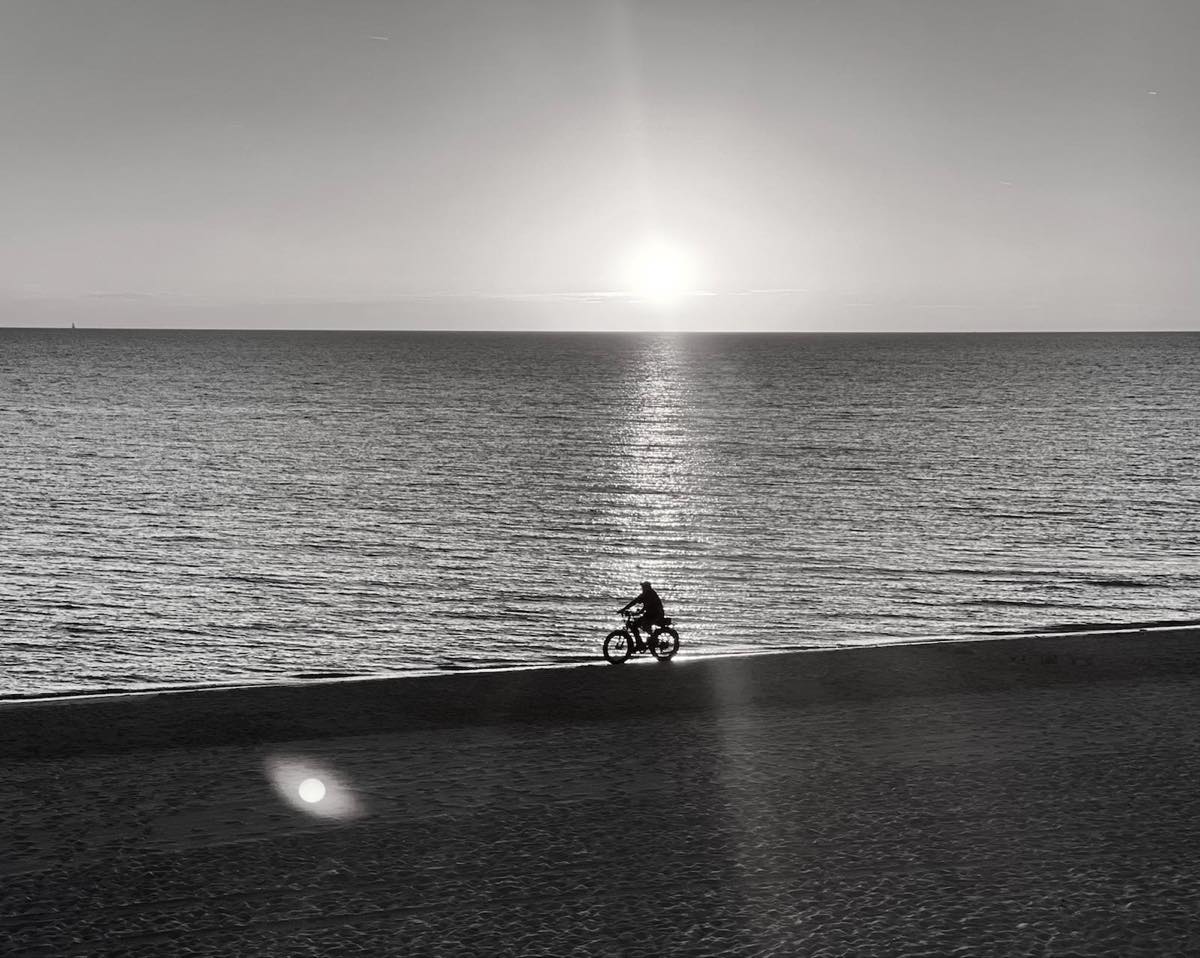 bikerumor pic of the day a photo of a cyclist on the beach taken from afar as the sun sets on the horizon, there is a glare in the camera lens and the photo seems to be in black and white.