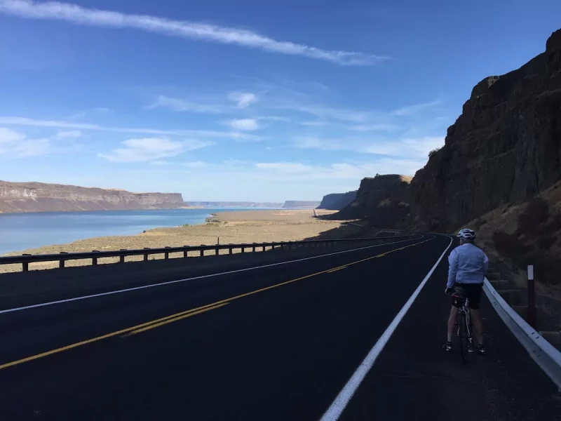 bikerumor pic of the day a cyclist is on the side of a road that follows a large river the mountain is shading the road but the sky is bright and the sun is shining on the river.