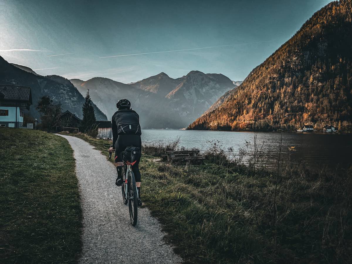 bikerumor pic of the day a gravel cyclist is on a narrow gravel path alongside a river with mountains in the distance.